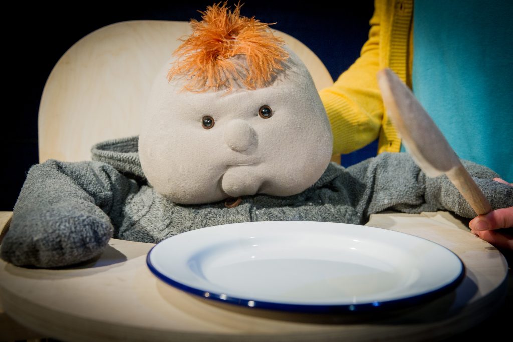 Beasty Baby puppet has a tuft of orange hair and wears a grey woollen outfit. He sits in a high chair with a white metal plate in front of him. The hands of a puppeteer can be seen manipulating him and in her other hand is a knife.