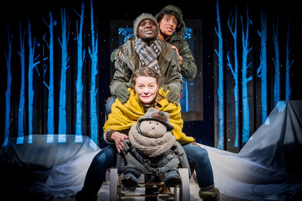 Beasty Baby puppet sits at the front of a wooden sleigh, a puppeteer sits behind him wrapped in a yellow woollen shawl and standing at the back of the sledge are two performers dressed in outdoor winter coats, hats and scarves. Behind them is a backdrop of blue illuminated trees.