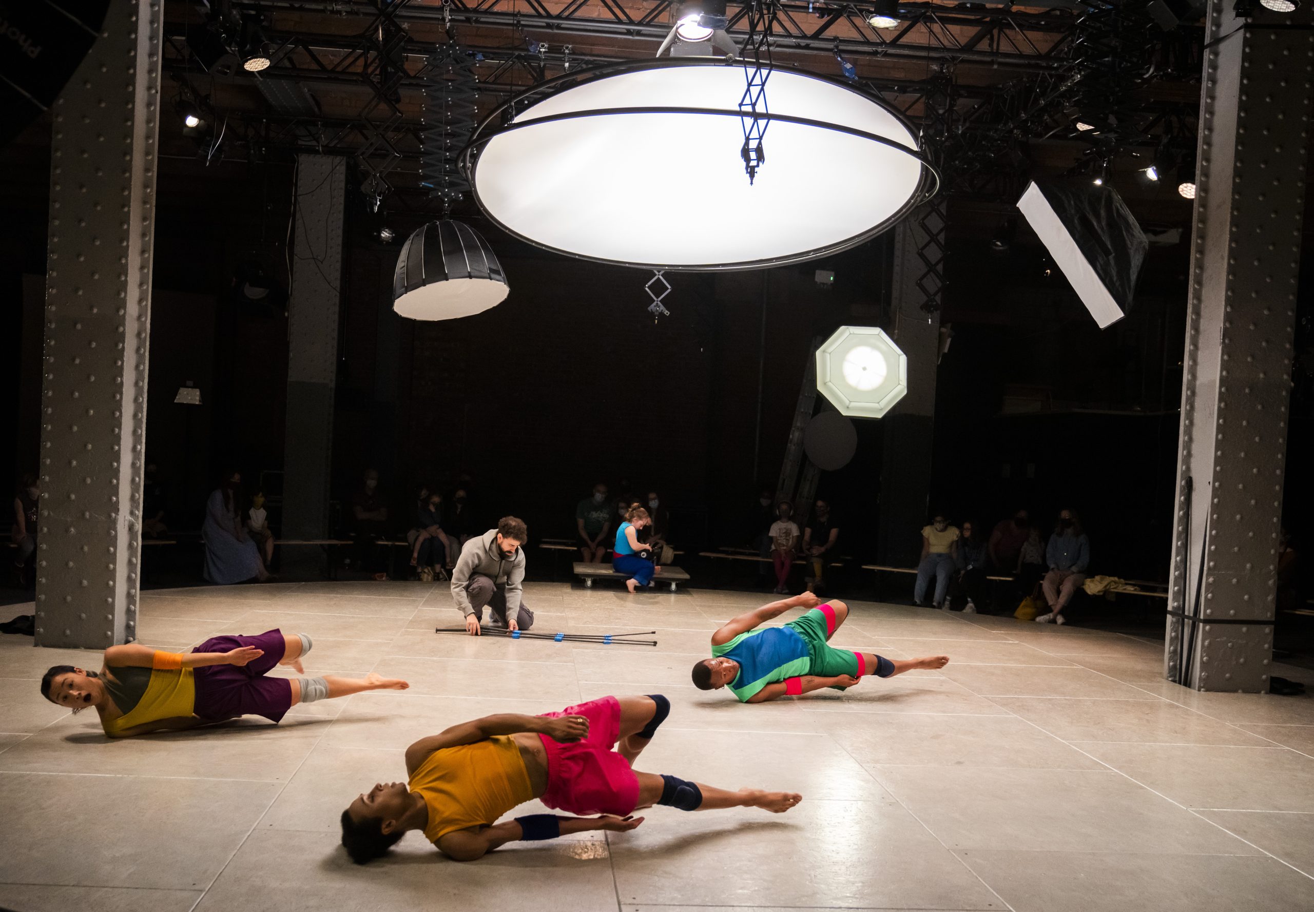 Three performers lie on stage. A fourth performer manipulates a camera tripod. The performers mimic the tripod's position with their bodies. Circle light reflectors are suspended above them.
