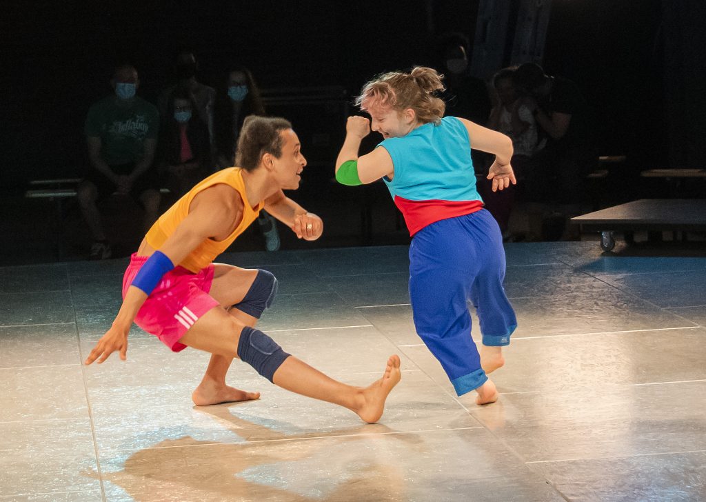 Two performers are on stage. One is crouched on the floor with one leg straight and the other runs next to them. They look at each other with upbeat facial expressions.