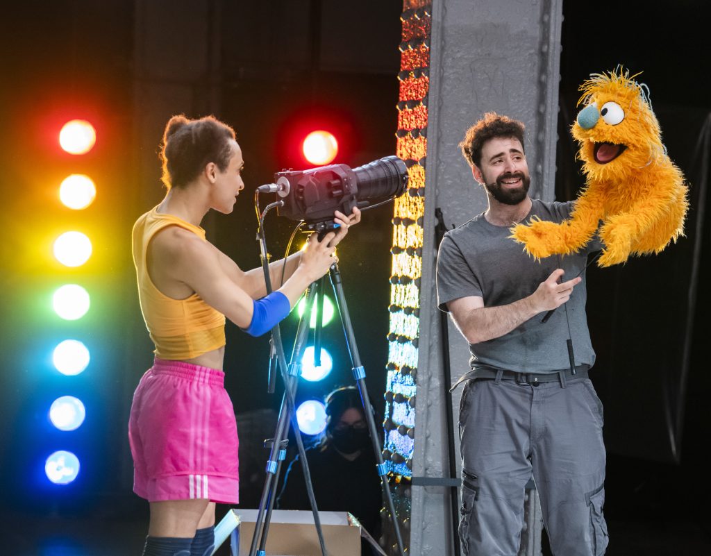 Two performers on stage. One is holding a camera aimed at the other. The other performer holds a muppet style orange puppet. There are multicoloured circle lights in the background.