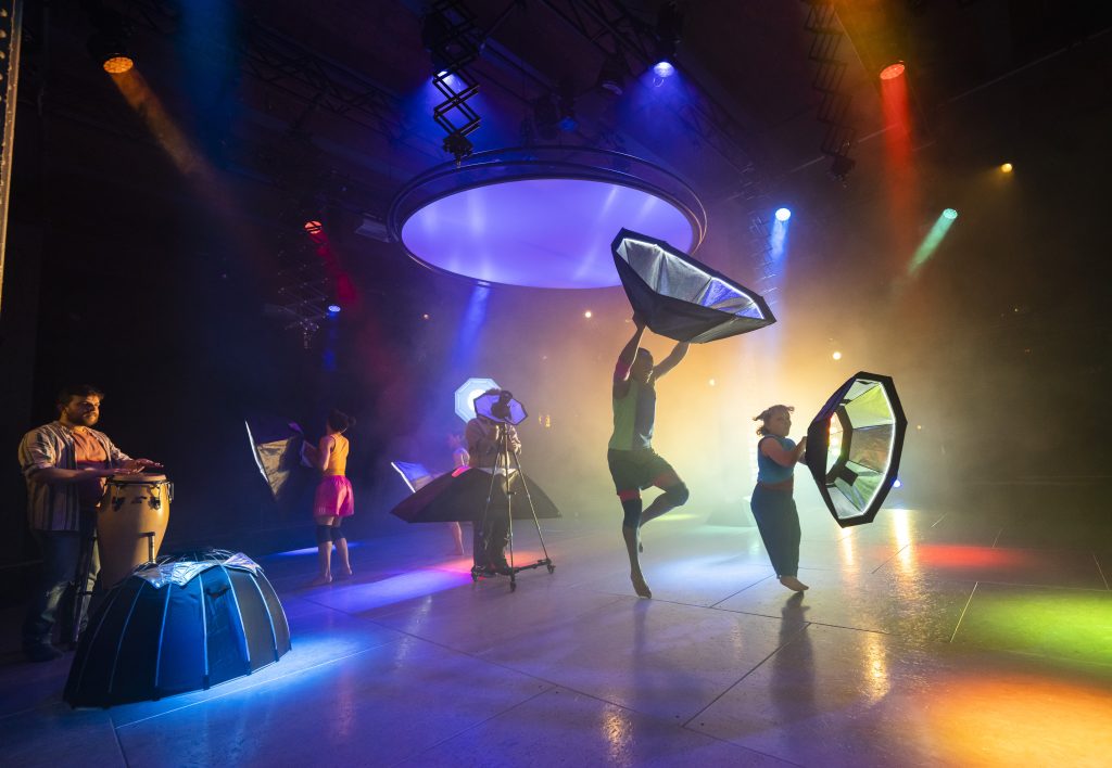 5 performers on stage. There are multicoloured spotlights. 4 of the performers dance in the space holding big film reflectors. The 5th performer on the left drums on a standing drum.