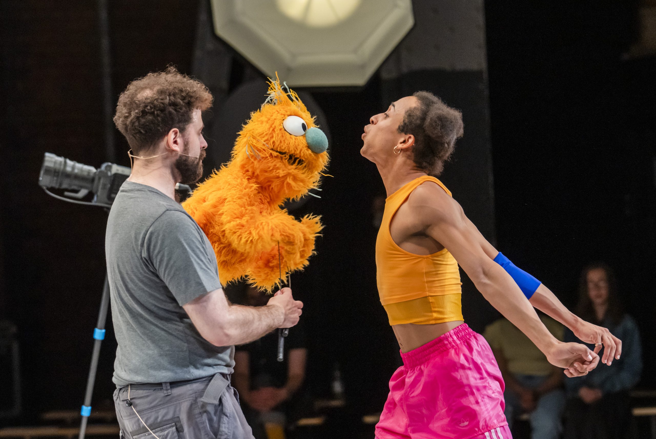 Two performers on stage. One is holding an orange furry muppet style puppet. The other performer blows air into the puppet's fur.