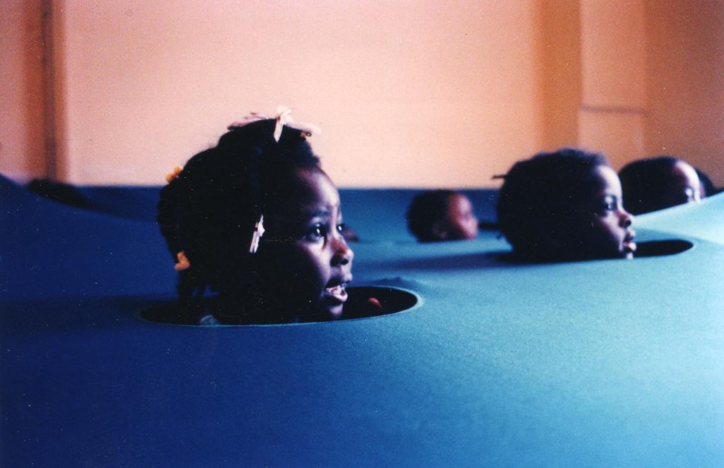4 young children pop their heads through holes cut from a piece of blue fabric, resembling the sea
