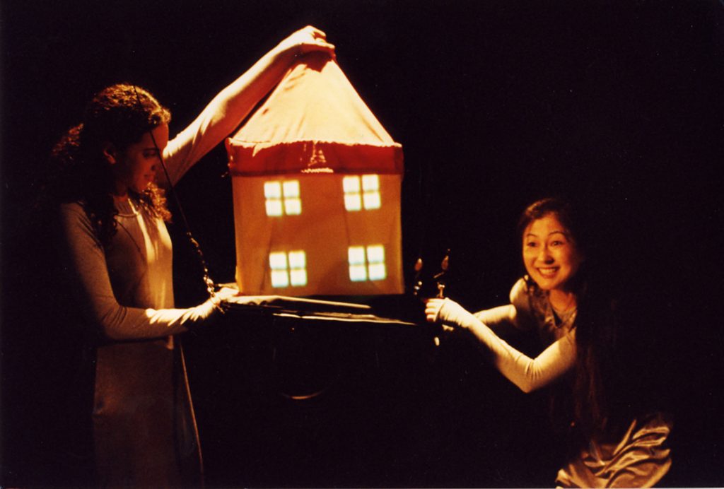 Two performers hold up a small house made of fabric between them. One of them is holding the house up by the top of its roof. The house is orange and it has four illuminated windows on the front.