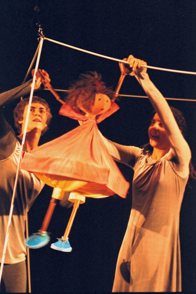 The girl puppet Belle, with an orange dress and blue shoes, is playing a game and swinging from a string structure. Belle is helped by two performers, who are enjoying the game with her.
