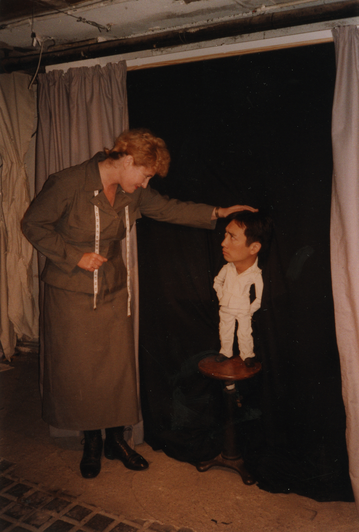 A performer wearing a matching jacket and skirt and a tape measure around her neck stands beside a humanette puppet: a small puppet body with a performer's head standing on top of a stool in front of a black screen.