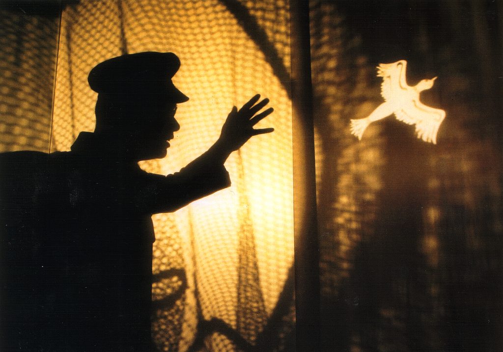 a human-like silhouette wearing a cap with arm outstretched towards an illuminated shape of a bird. There is netting effect in the background lit from behind