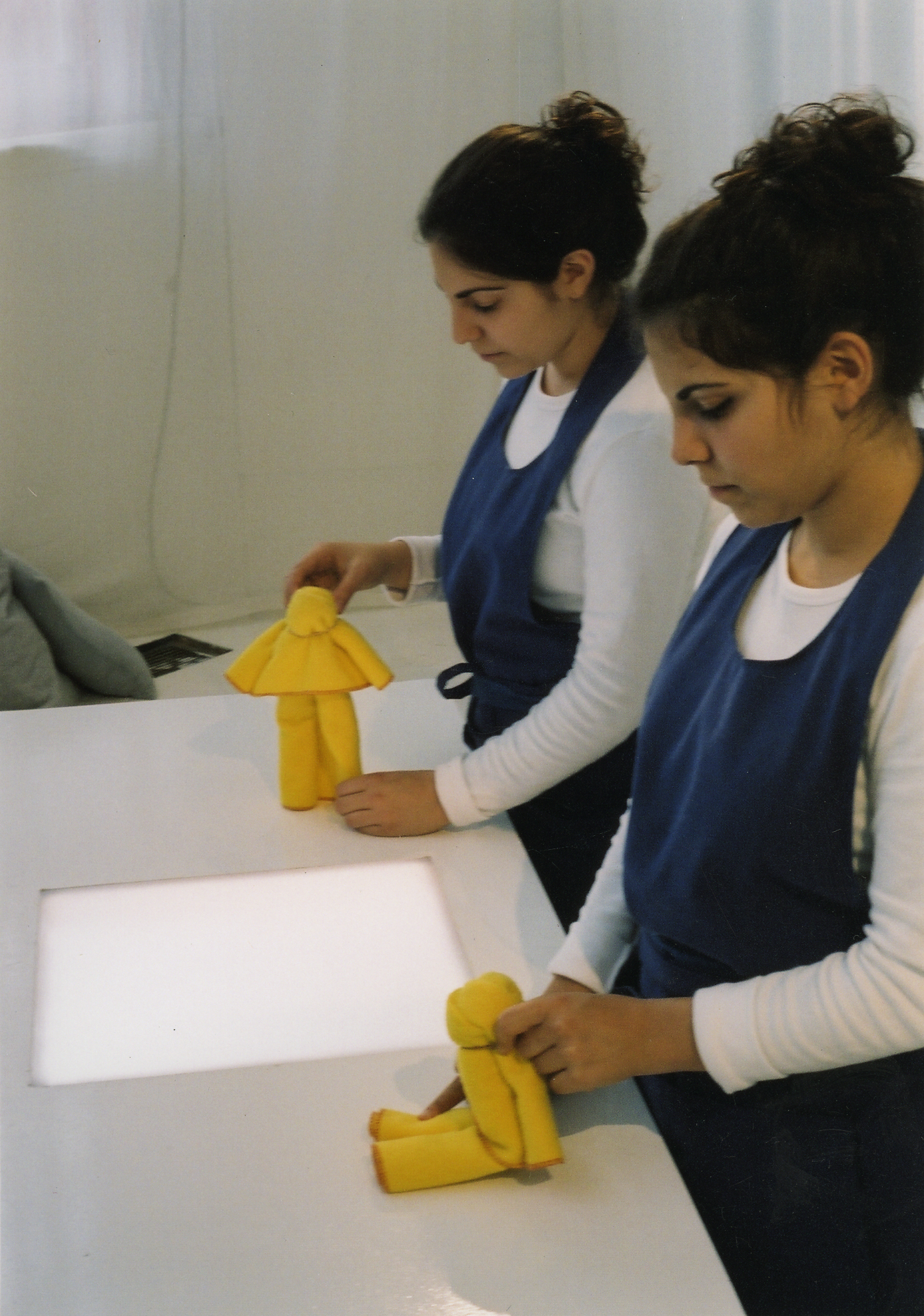 2 performers wearing blue aprons stand at a counter top each manipulating a small figure made out of a yellow duster.