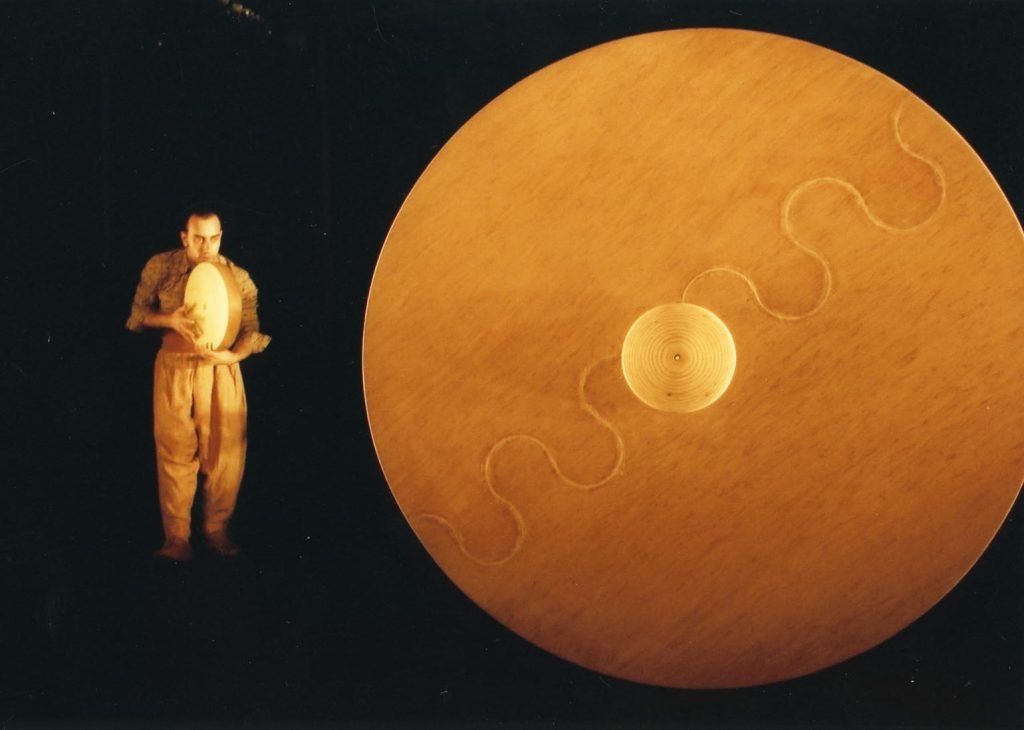 a male performer stands playing a tambourine-shaped drum. The shape of the instrument reflects a much larger circular wooden object next to the performer, which has a wavy line across the length of it and a smaller circle in the middle.