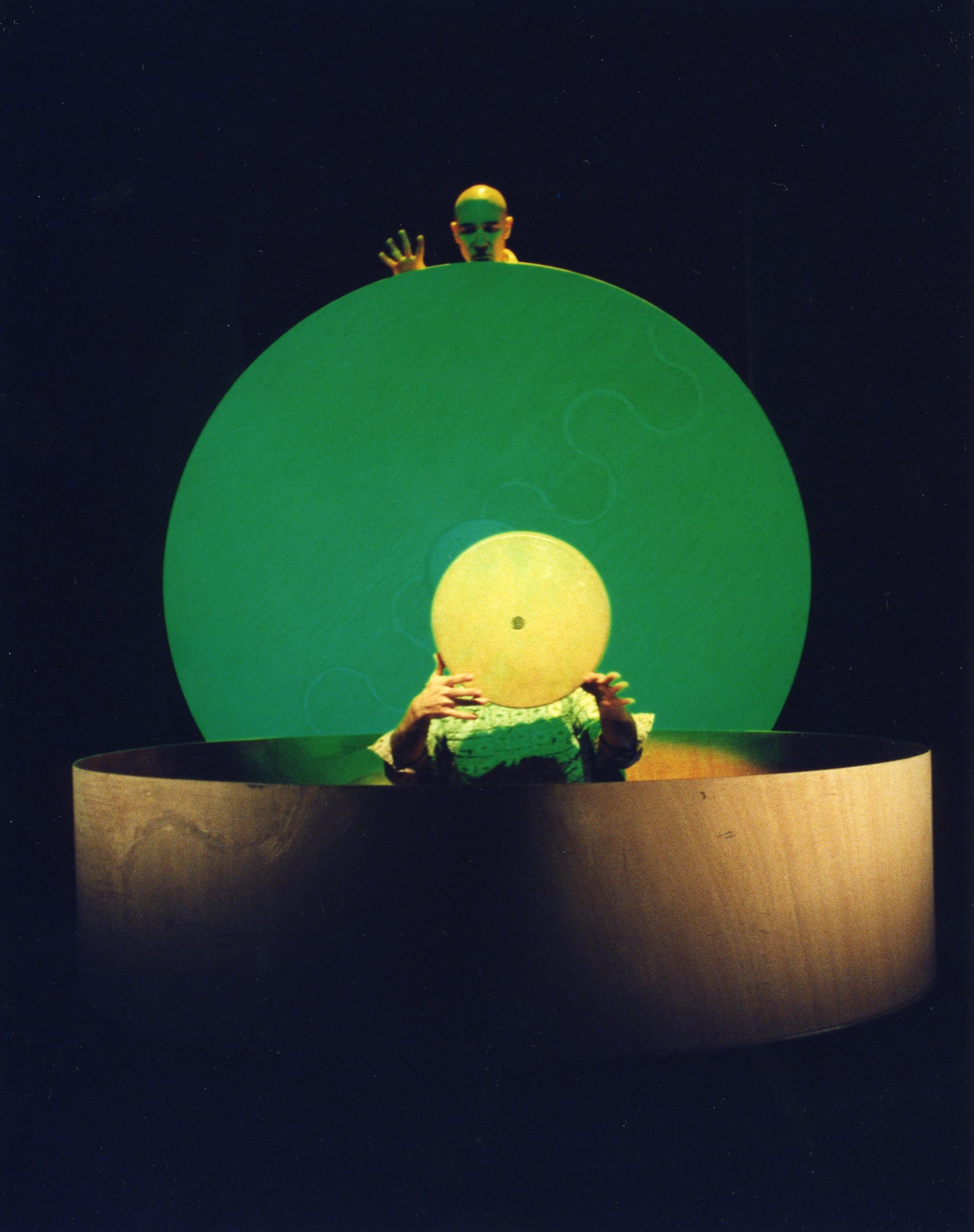 In the background, a performer looks over the top of a large green circular object. His head and one hand are visible. He looks down at another performer who sits in the middle of a large circular wooden shape holding a small circular shape in front of their head.