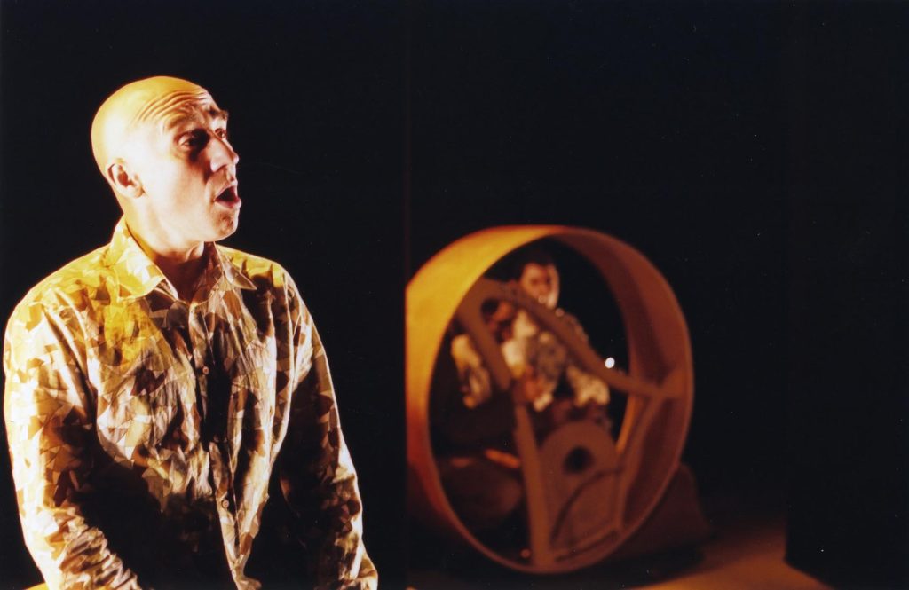 in the foreground in focus a male performer sings while in the background, out of focus, another male performer crouches behind a circular harp and plays it.