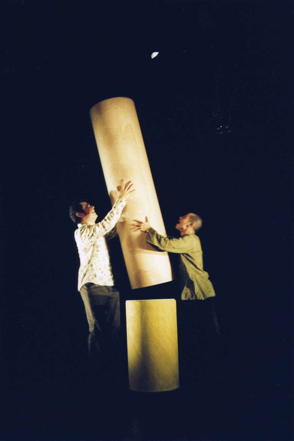 2 male performers face each other each with their hands on a large cylinder shaped object which they hold between them. A separate cylinder shaped object is on the floor below.