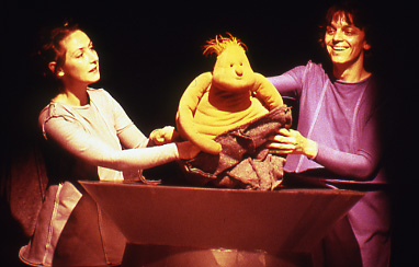 an orange puppet baby with a tuft of orange hair sits on a piece of red fabric and is manipulated by 2 puppeteers either side of it.