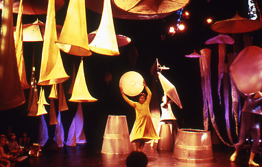 a stage set which has a number of yellow fabric cones and purple/red coloured pieces of fabric hanging from the ceiling. 3 performers hold fabric shapes above their heads and a young children in the audience watch from the side.