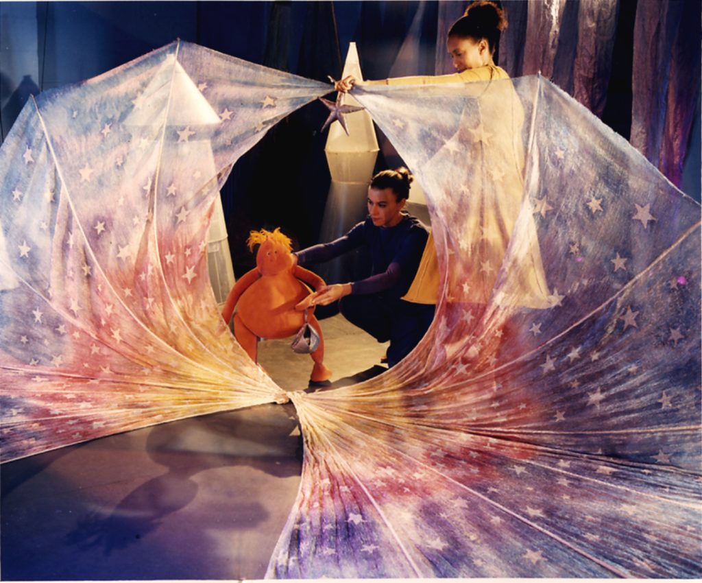 a small orange coloured puppet with a tuft of orange hair stands on the floor manipulated by a puppeteer. A second puppeteer stands next to them holding a large transparent piece of fabric covered in stars which encircles the puppet.