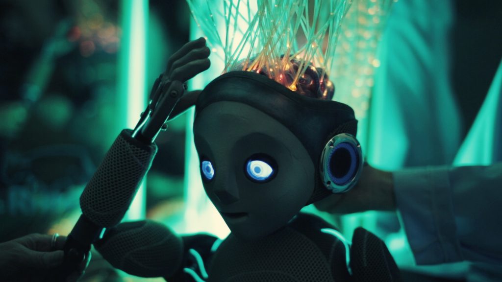 Robot Boy puppet raises his hand towards his brain which has wires attached which are lit from within his brain.