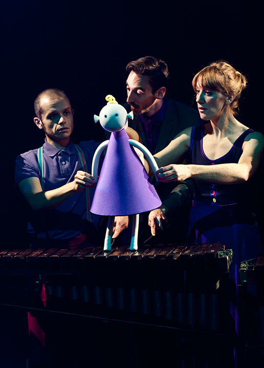 A conical shaped puppet girl wearing a purple dress stands on a xylophone as 3 performers manipulate her and her feet play a tune.