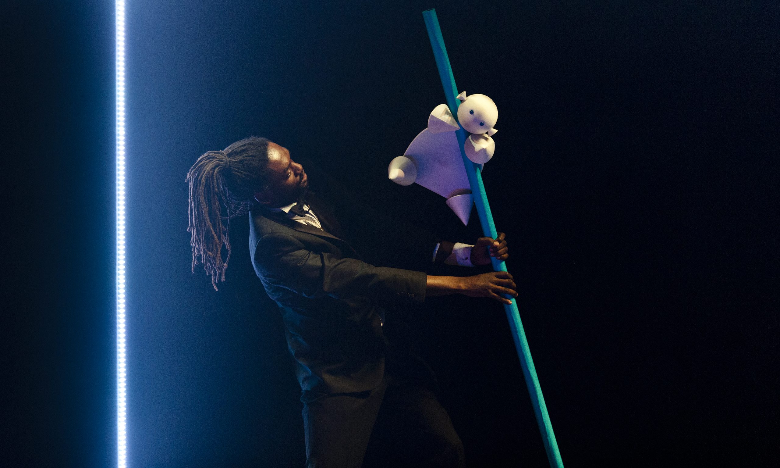 A performer with dreadlocks holds a blue pole at an angle as a puppet baby slides down it.