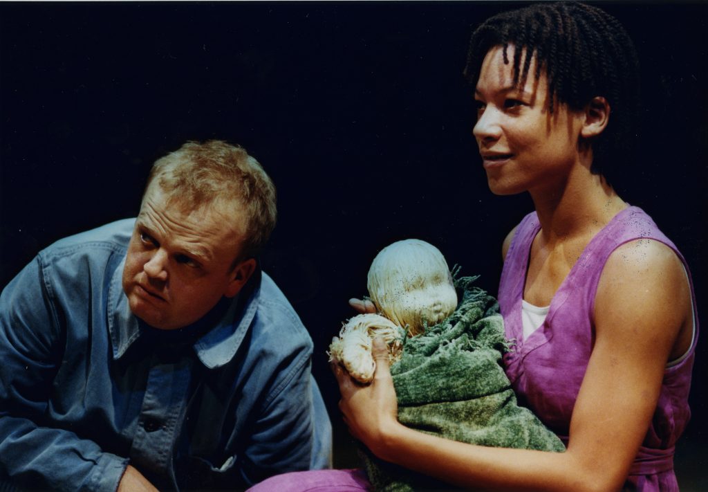 An image from the early 1990s. On the left of the image actress Nina Sosanya with short braided hair cradles a wooden baby puppet wrapped in fabric. On the right actor Toby Jones leans looking to the side.