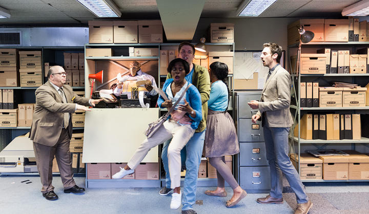 Four angry looking performers crowd around a filing cabinet in which sits a puppet who holds paper receipts. The performer who plays the Bank Manager in the show is trying to calm them all down.