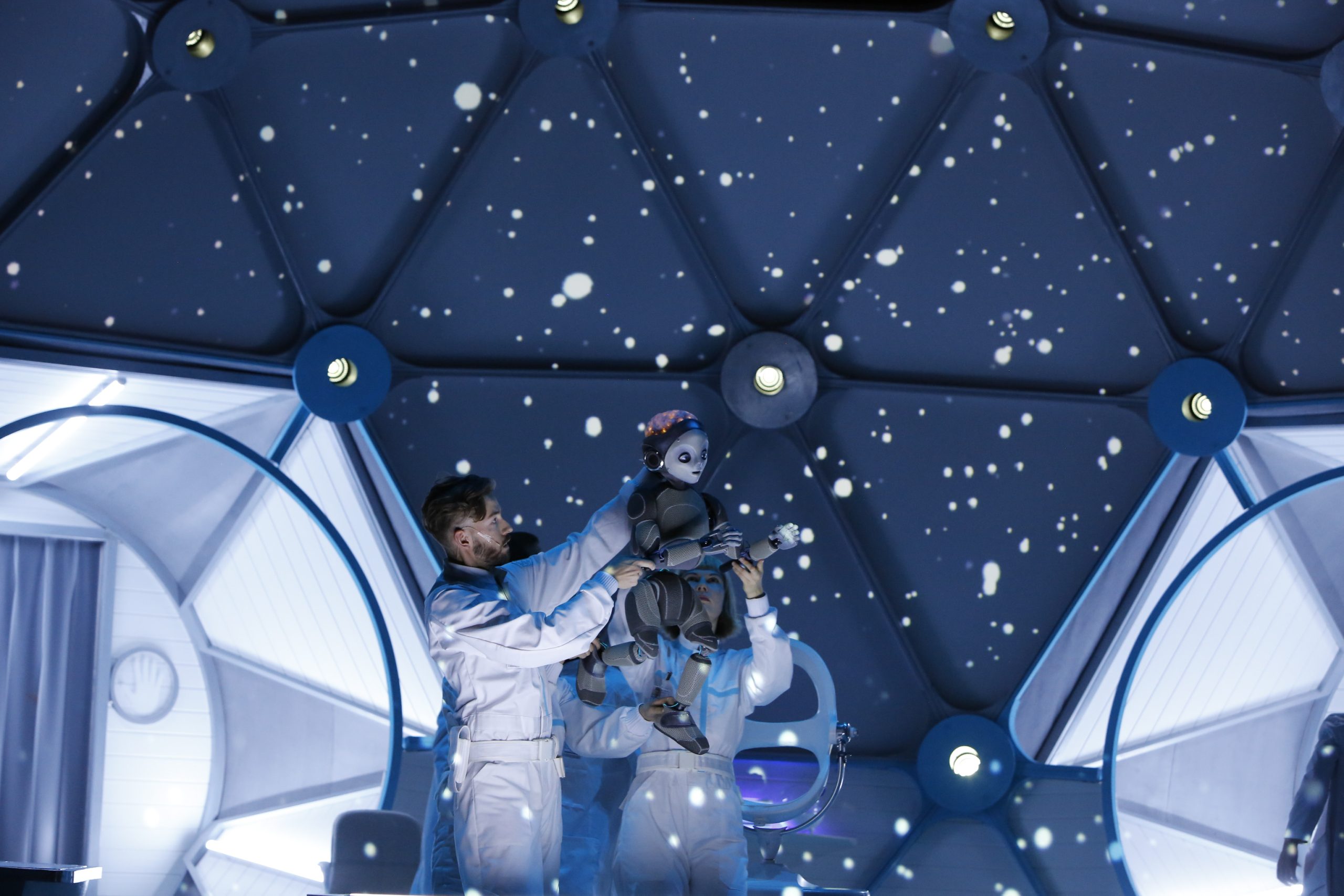 Two puppeteers manipulate a Robot Boy puppet across the stage at head height. The backdrop digital image gives the impression of stars in the sky.
