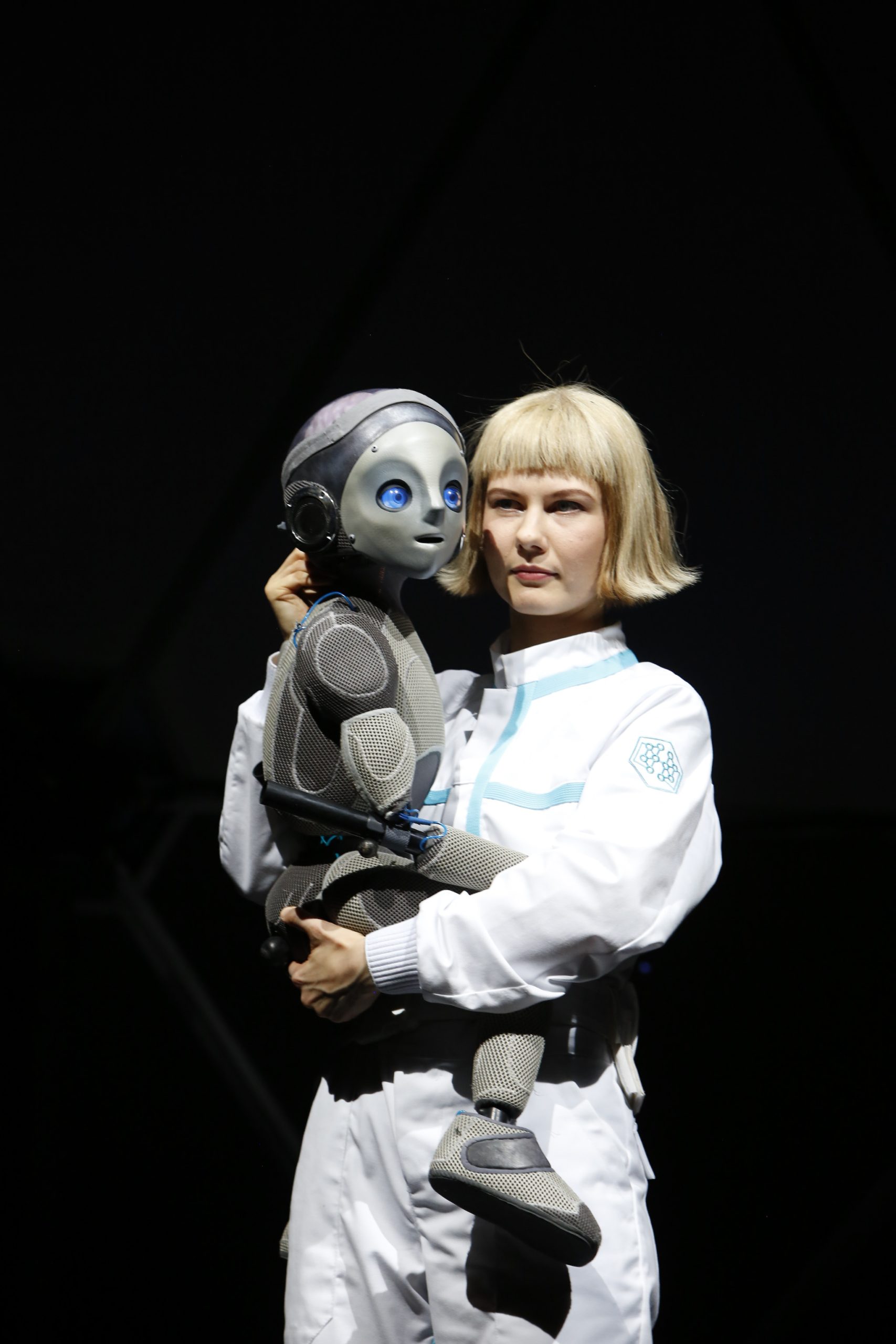 a puppeteer manipulate Robot Boy puppet who she holds against her waist against a black background.