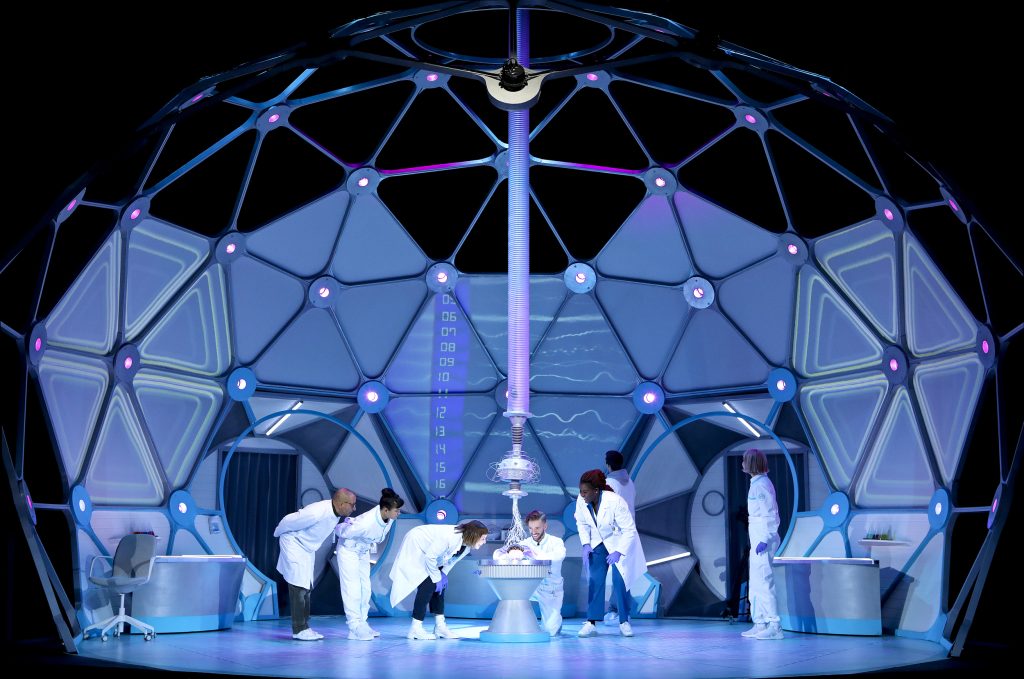 A stage set science lab made of of triangular segments in the shape of a head. There are 7 performers on stage all dressed in white lab coats or boiler suits. Some stand and others bend over looking at a brain on a stand. A long cylindrical tube hangs from the ceiling with wires at the lowest end attached to the brain.