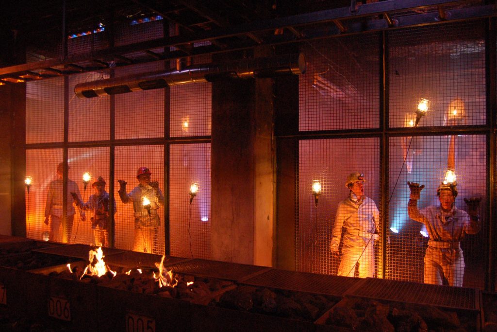 5 performers dressed in pale overalls with hardhats sing from behind a wire mesh screen. They are lit by a number of industrial style lights. In the foreground are crates full of coal which are alight.