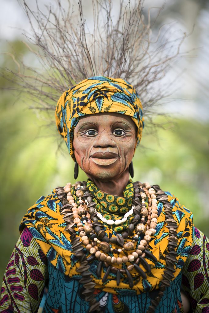 A puppet of a black woman in traditional South African clothing in purple, blue and yellow colours. She wears layers of wooden beads around her neck and has a yellow headscarf on with long twigs for hair. She looks into camera.