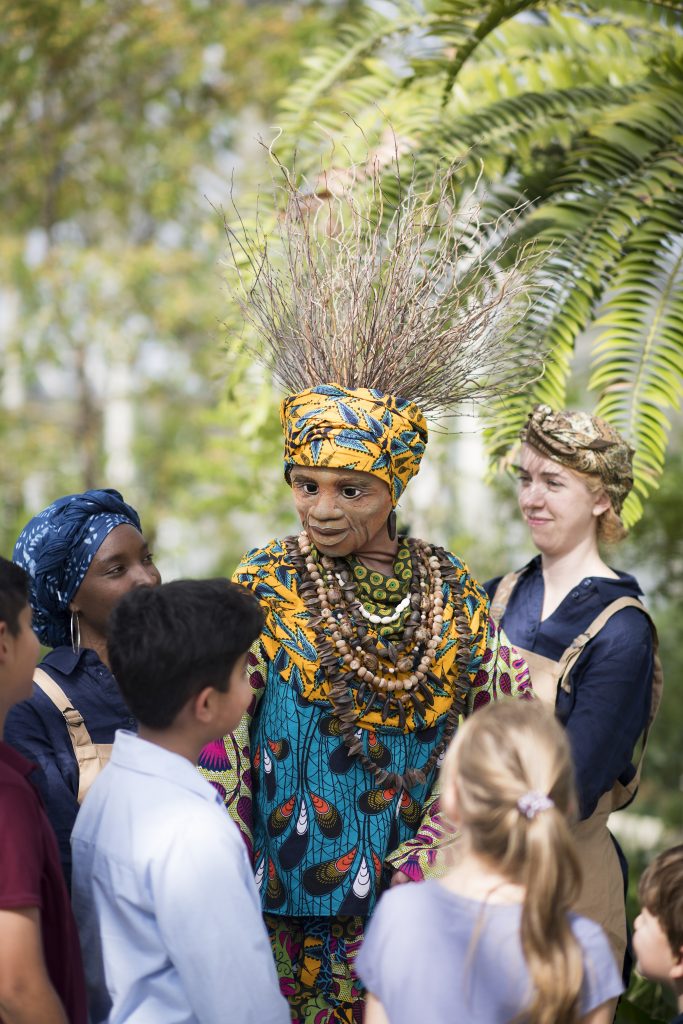 A puppet of a black woman in traditional South African clothing in purple, blue and yellow colours. She wears layers of wooden beads around her neck and has a yellow headscarf on with long twigs for hair. She is puppeteered by two performers in blue overalls and headscarfs. They are among green leaves and trees. There is an audience of children looking at the puppet.