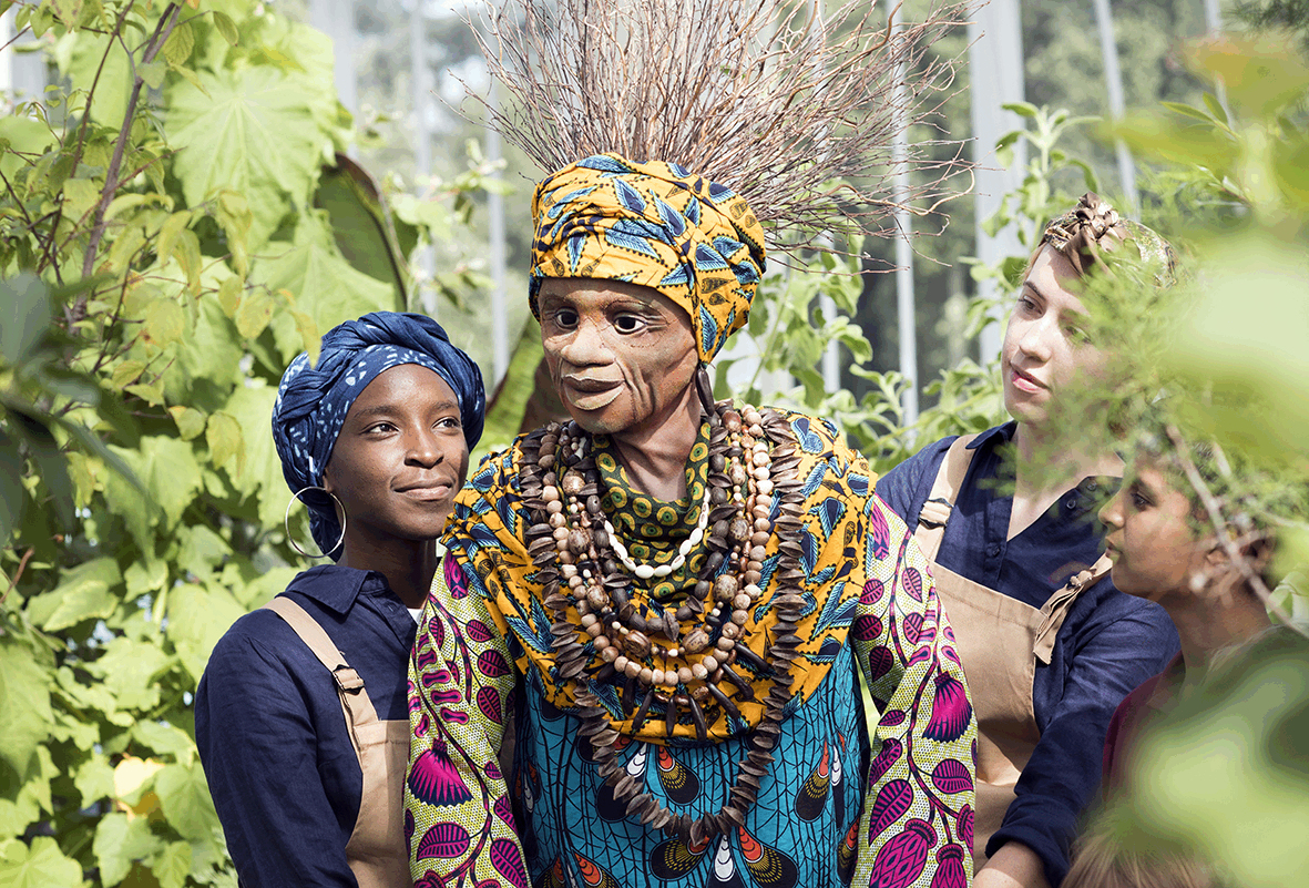 A puppet of a black woman in traditional South African clothing in purple, blue and yellow colours. She wears layers of wooden beads around her neck and has a yellow headscarf on with long twigs for hair. She is puppeteered by two performers in blue overalls and headscarfs. They are among green leaves and trees.
