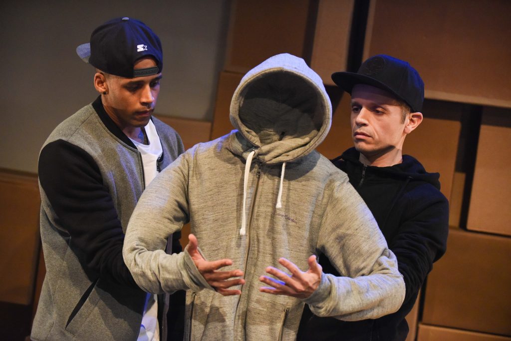 A puppet in a grey zip up hoodie is manipulated by man in black jacket and a man in a black hat.