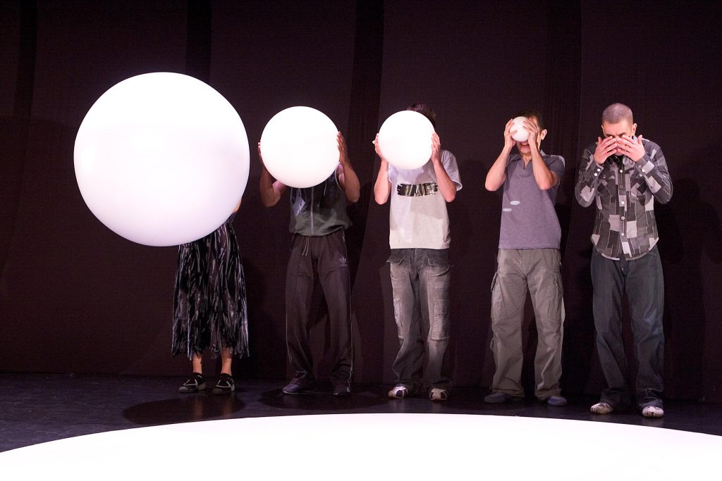 Five friends stand on the edge of a white circle. The man on the right holds his hands over his eyes and the remaining four hold white balls which gradually increase in size until the last person is almost completely covered by a huge ball.