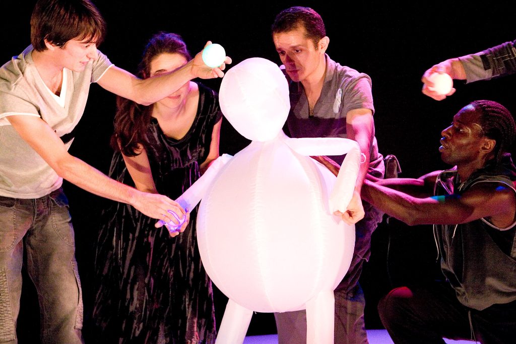 Four performers manipulate the movements of a boy made out of white balloons, trying to encourage him to play a ball game.
