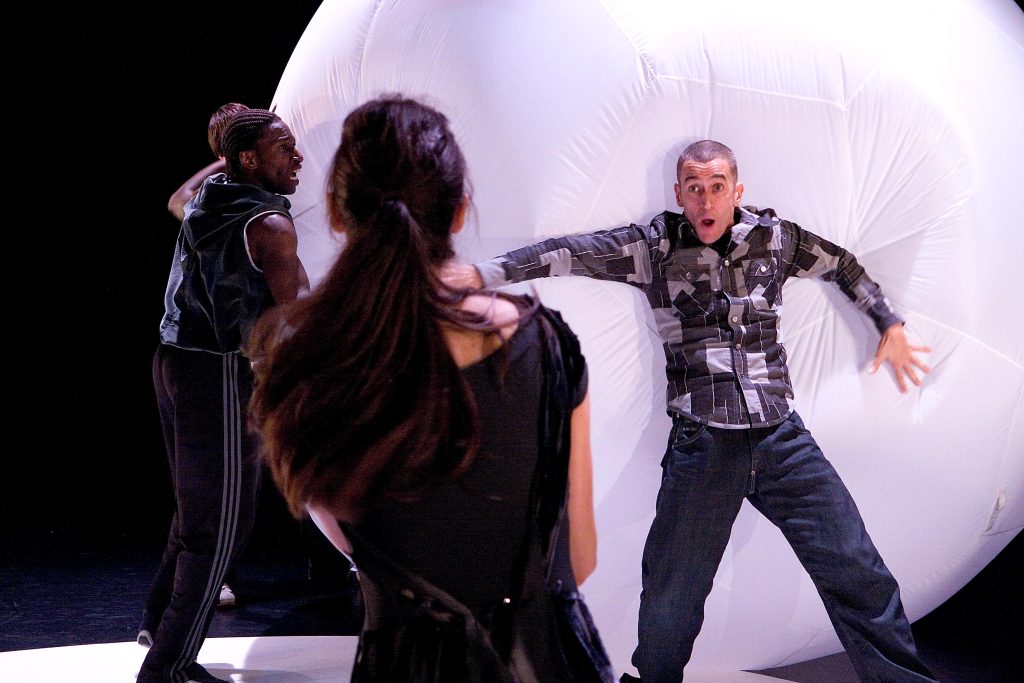 A man falls back with his arms stretched out onto a giant balloon as if the wind has pushed him. He looks shocked as a woman watches him and a man tries to hold on to the giant balloon.