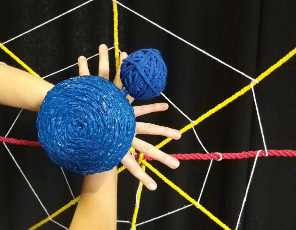 2 hands crossed in front of a spiders web made from yellow, red and white string. Two balls made from blue string