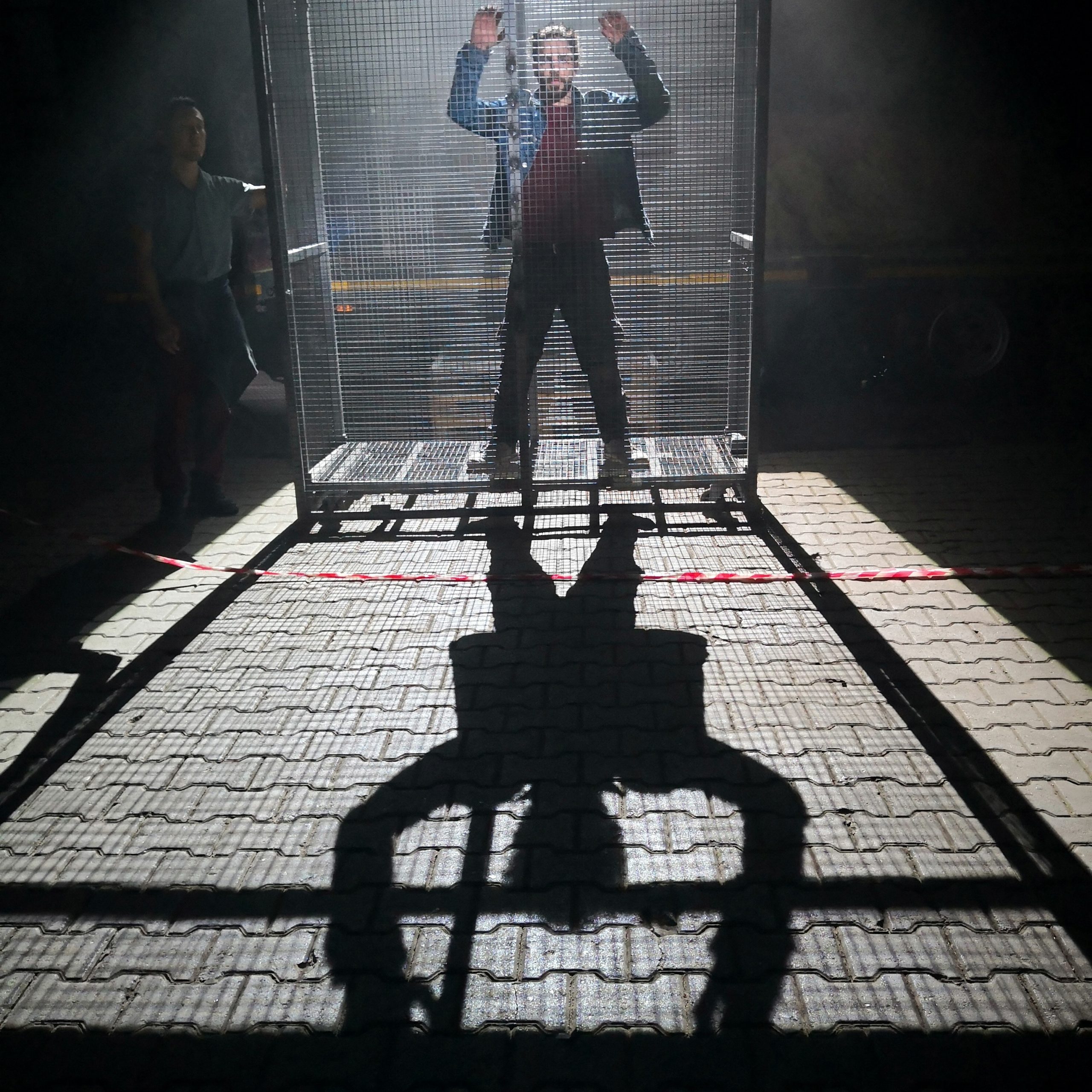a performer stands in a cage with his arms raised. He is lit from behind and his body casts a long shadow in the foreground.