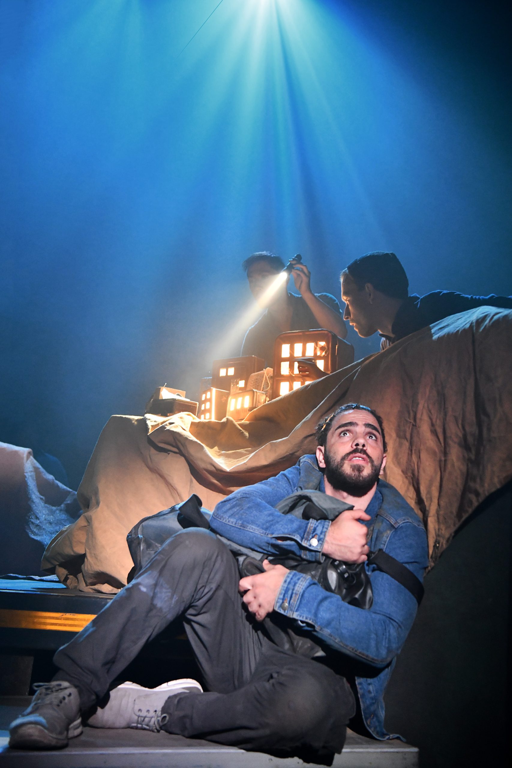 a performer sits on the ground clutching his travel bag. Behind him another performer holds a torch shining a light on a miniature cityscape and a third performer looks on.
