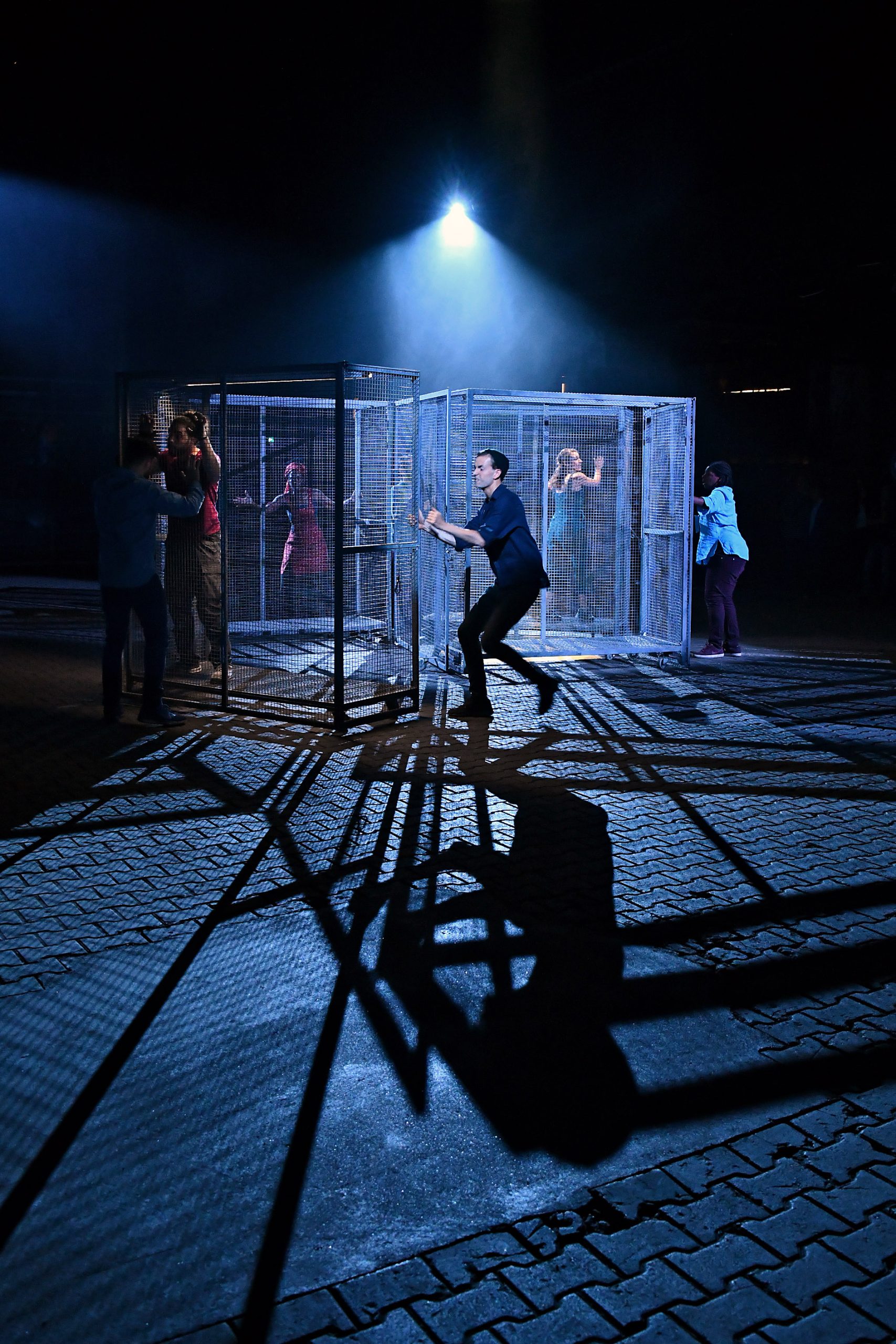 a group of performers are moving large metal cages in a darkened space with a spotlight from above. Some performers are inside the cages.