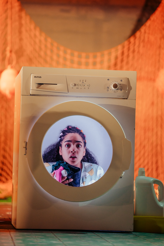 A white washing machine sits on-stage with a young woman inside it. Her shocked face is visible through the round transparent door. In the background there is hanging orange netting and to the right of the machine is a green detergent bottle.