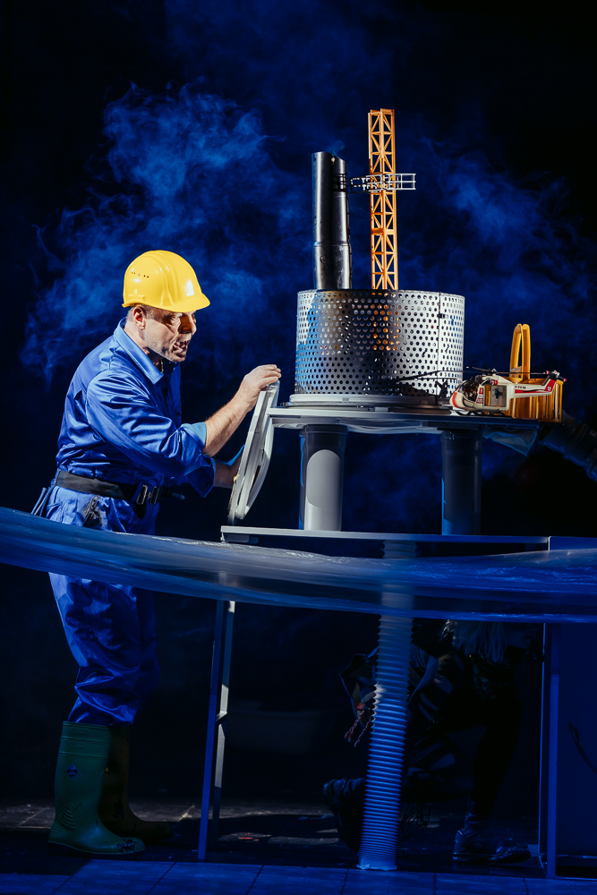 A man with in blue satin overalls and a yellow hardhat is on stage. He is looking at and holding with one hand a large pile of objects set up to look like an oil rig. The objects are made of metal.
