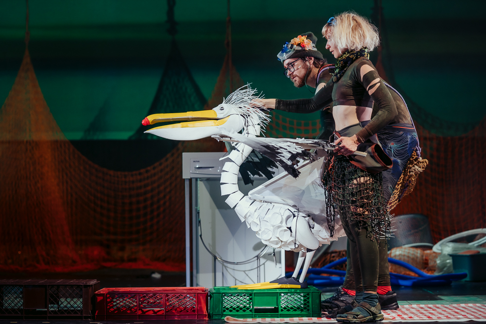 Two performers in green torn combat clothes puppeteer a white pelican made of plastic cups. the pelican has a yellow beak and red nose tip made of a bottle cap. The pelican is standing on plastic crates on a stage.