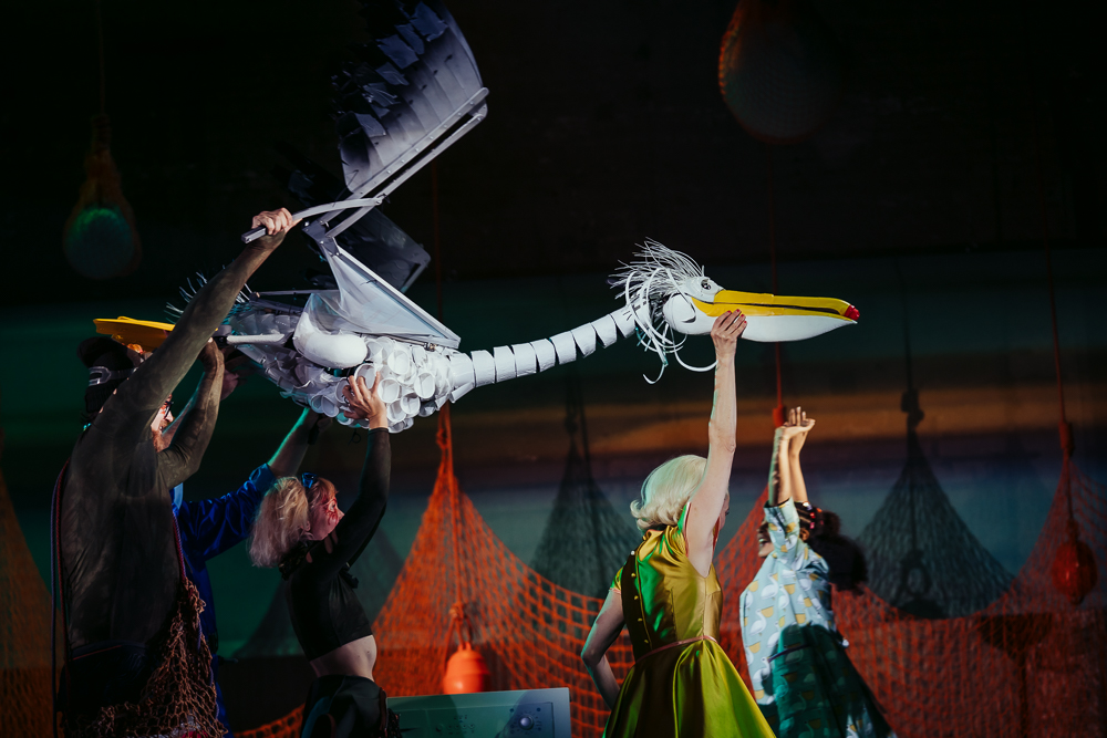 A white puppet pelican made of plastic cups is puppeteered by 3 performers to look as though it is flying on stage.