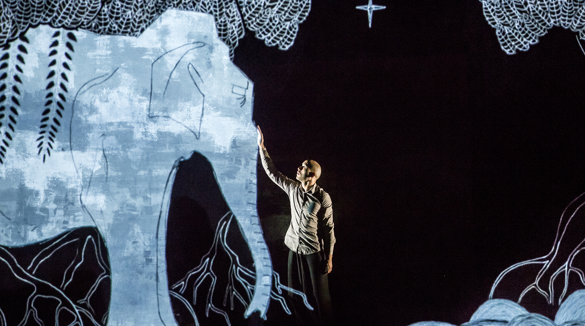 Against a black screen we see a blue drawn elephant and shrubs projected. Standing touching the elephant's trunk is a dancer wearing black trousers and a light linen shirt.