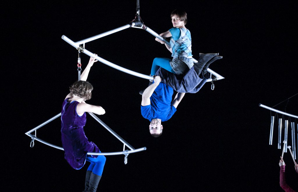 3 aerialist performers hang from sections of a metal mobile which is suspended from above. In the bottom right corner, the arms of a musician are seen holding musical mallets playing a metal tubular instrument.
