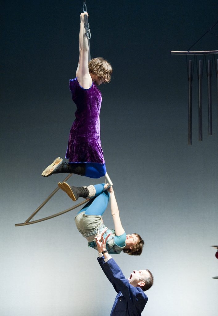 2 aerial performers are hanging from a metal mobile suspended from above while a third performer on the ground holds his arms up with an alarmed expression in case the aerialists should fall.