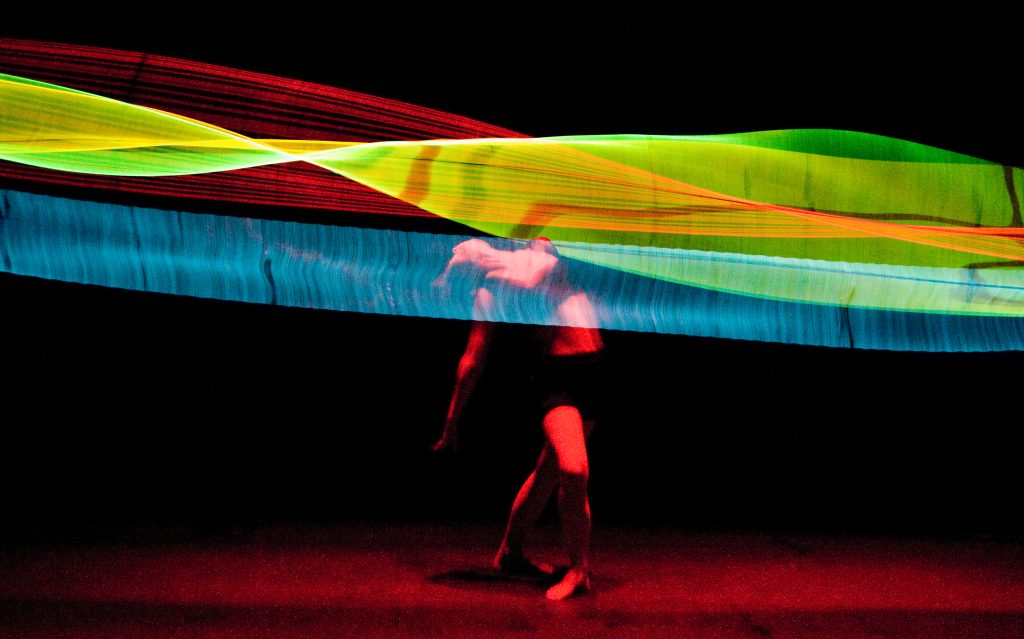 a performer dances beneath strips of different coloured lights which are moving to create an abstract wave effect.