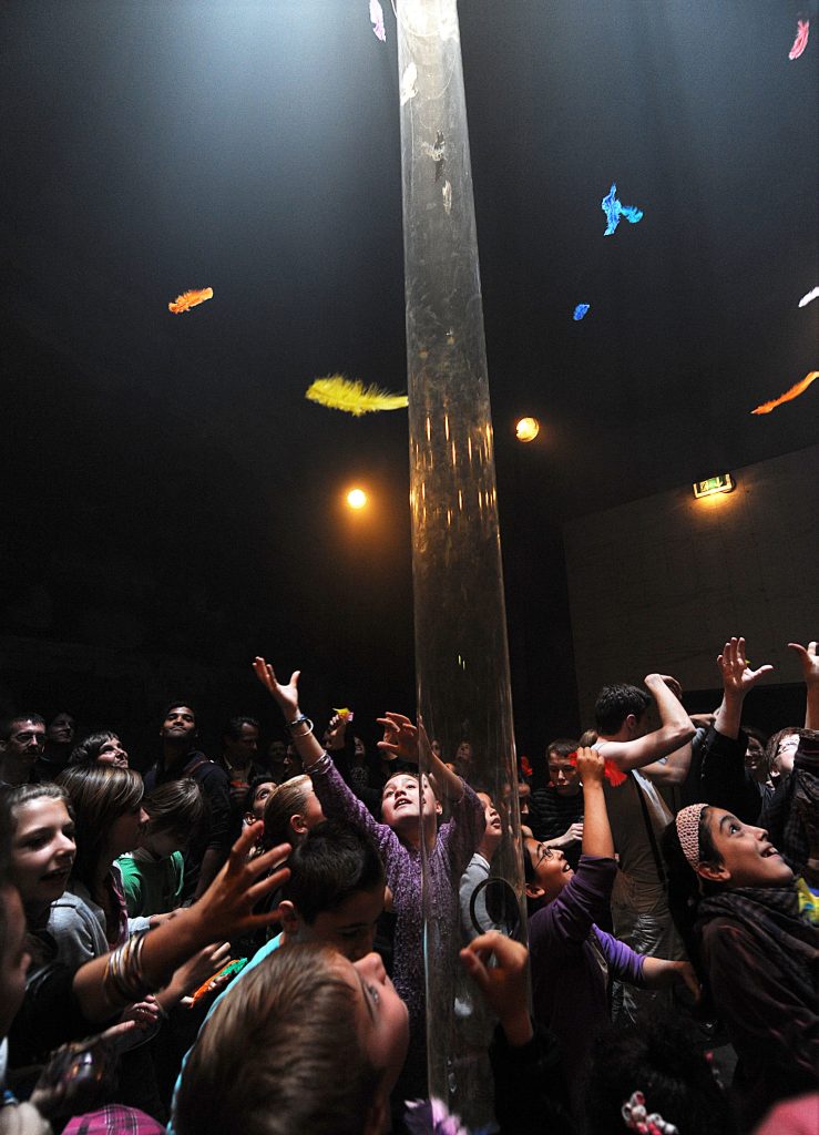 children reach for coloured feathers as they fall from above. A tall cylindrical plastic tube hangs from the ceiling.