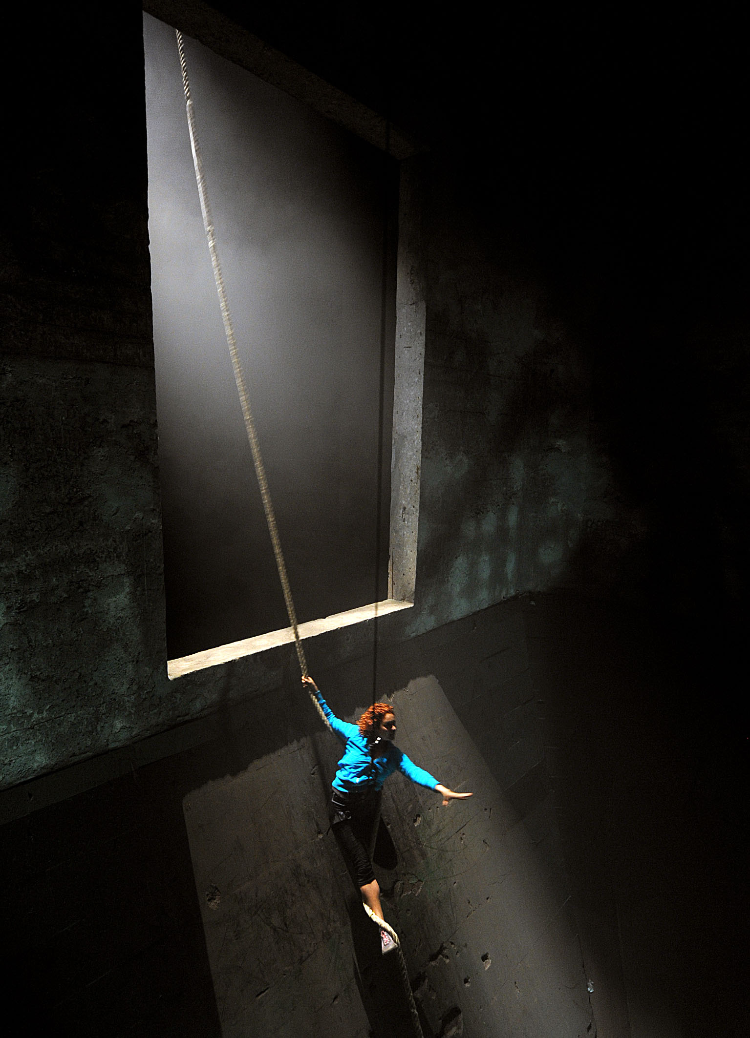 a performer in a blue top and black shorts dangles from a rope with one hand. The rope is hanging from the top of a long concrete shute.